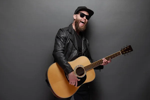 Gitárjátékos énekel a zenei stúdióban. Hipster gitáros szakállal és fekete ruhát játszik az akusztikus gitár — Stock Fotó