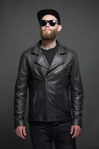 Uomo con giacca biker in pelle o giacca asimmetrica con zip e cappuccio nero, jeans e occhiali da sole. Bello hipster uomo su sfondo grigio — Foto Stock