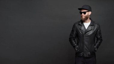Siyah şapkalı, kot pantolonlu ve güneş gözlüklü deri bisikletçi ceketi veya asimetrik zip ceket giyen adam. Kopya alanı ile gri arka plan üzerinde Yakışıklı hipster adam