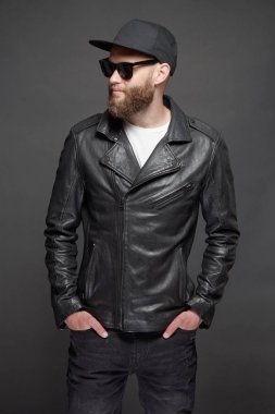 Siyah şapkalı, kot pantolonlu ve güneş gözlüklü deri bisikletçi ceketi veya asimetrik zip ceket giyen adam. Gri arka plan üzerinde Yakışıklı hipster adam