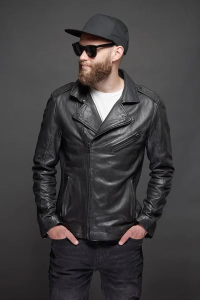 Man draagt lederen biker jasje of asymmetrische zip jas met zwarte pet, jeans en zonnebril. Knappe hipster man over grijze achtergrond — Stockfoto