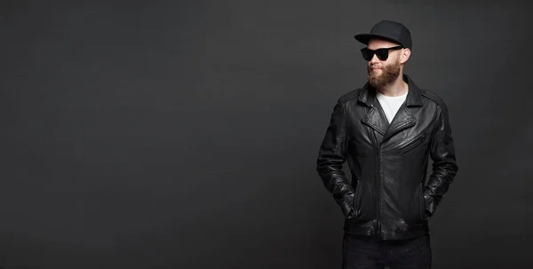 Мужчина в кожаной куртке байкера или асимметричной куртке с черной кепкой, джинсами и солнцезащитными очками. Красивый хипстер на сером фоне с копировальным пространством — стоковое фото