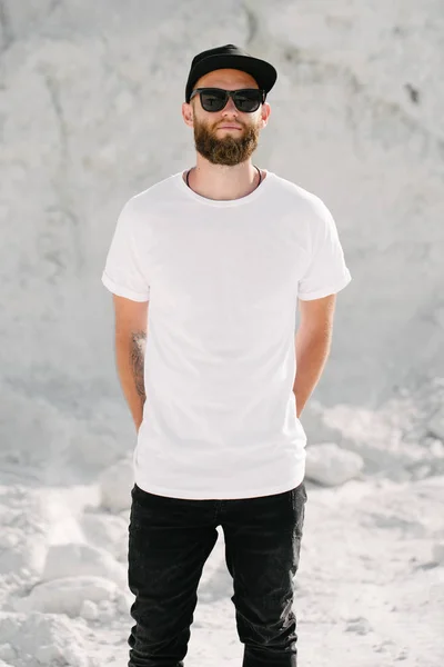 Hipster tampan model laki-laki dengan jenggot mengenakan putih kosong t-shirt dan topi bisbol dengan ruang untuk logo atau desain dalam gaya perkotaan biasa — Stok Foto