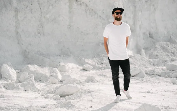 Hipster guapo modelo masculino con barba vistiendo camiseta blanca en blanco y una gorra de béisbol con espacio para su logotipo o diseño en estilo urbano casual —  Fotos de Stock