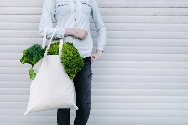 Woman holding an eco bag filled with grocery. Vegetables and fruits are hanging from a bag. Ecology or environment protection concept. White eco bag for mock up.