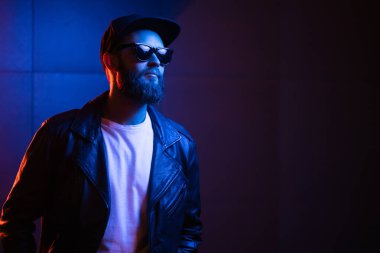 Şehir sokaklarında hipster yakışıklı adam Neon işaretler tarafından aydınlatılmış olmak. O siyah kap, kot pantolon ve güneş gözlüğü ile deri motosikletçi ceket veya asimetrik fermuar ceket giyiyor.