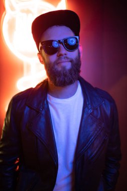 Şehir sokaklarında hipster yakışıklı adam Neon işaretler tarafından aydınlatılmış olmak. O siyah kap, kot pantolon ve güneş gözlüğü ile deri motosikletçi ceket veya asimetrik fermuar ceket giyiyor.
