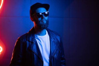 Şehir sokaklarında hipster yakışıklı adam Neon işaretler tarafından aydınlatılmış olmak. O siyah kap, kot pantolon ve güneş gözlüğü ile deri motosikletçi ceket veya asimetrik fermuar ceket giyiyor.