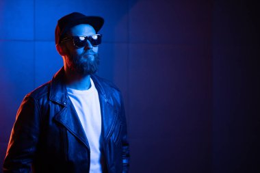 Şehir sokaklarında hipster yakışıklı adam Neon işaretler tarafından aydınlatılmış olmak. O siyah kap, kot pantolon ve güneş gözlüğü ile deri motosikletçi ceket veya asimetrik fermuar ceket giyiyor.