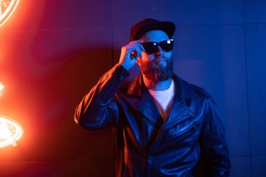 Şehir sokaklarında hipster yakışıklı adam Neon işaretler tarafından aydınlatılmış olmak. O siyah kap, kot pantolon ve güneş gözlüğü ile deri motosikletçi ceket veya asimetrik fermuar ceket giyiyor.