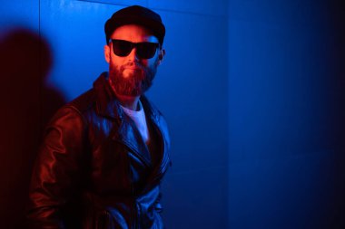 Şehir sokaklarında hipster yakışıklı adam Neon işaretler tarafından aydınlatılmış olmak. O siyah kap, kot pantolon ve güneş gözlüğü ile deri motosikletçi ceket veya asimetrik fermuar ceket giyiyor.