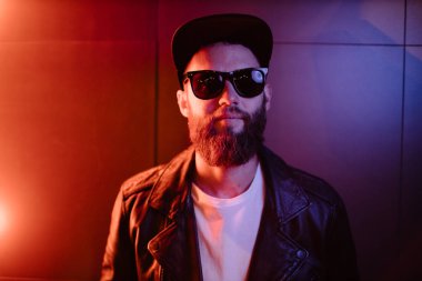 Şehir sokaklarında hipster yakışıklı adam Neon işaretler tarafından aydınlatılmış olmak. O siyah kap, kot pantolon ve güneş gözlüğü ile deri motosikletçi ceket veya asimetrik fermuar ceket giyiyor.