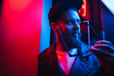 Şehir sokaklarında hipster yakışıklı adam Neon işaretler tarafından aydınlatılmış olmak. O siyah kap, kot pantolon ve güneş gözlüğü ile deri motosikletçi ceket veya asimetrik fermuar ceket giyiyor.