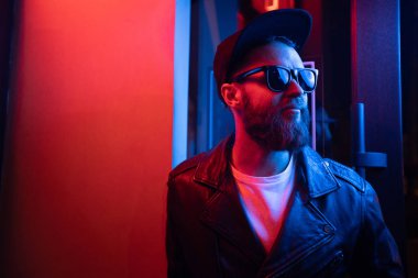 Şehir sokaklarında hipster yakışıklı adam Neon işaretler tarafından aydınlatılmış olmak. O siyah kap, kot pantolon ve güneş gözlüğü ile deri motosikletçi ceket veya asimetrik fermuar ceket giyiyor.