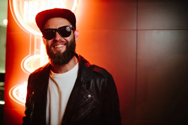 Hipster handsome man on the city streets being illuminated by neon signs. He is wearing leather biker jacket or asymmetric zip jacket with black cap, jeans and sunglasses. — Stock Photo, Image