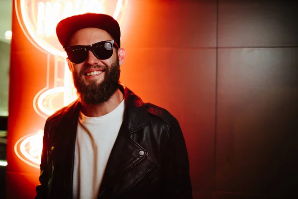 Hipster homem bonito nas ruas da cidade sendo iluminado por sinais de néon. Ele está vestindo jaqueta de couro motociclista ou casaco zip assimétrico com boné preto, jeans e óculos de sol . — Fotografia de Stock