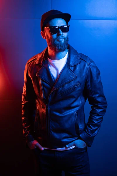 Hipster gutaussehender Mann auf den Straßen der Stadt, beleuchtet von Leuchtreklamen. er trägt Biker-Lederjacke oder asymmetrische Reißverschlussjacke mit schwarzer Mütze, Jeans und Sonnenbrille. — Stockfoto