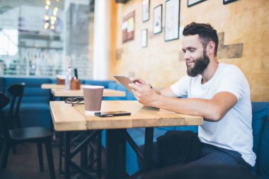 Bir restoranda ya da kahve civarında tablet bilgisayar kullanan adam. Beyaz tişört giyiyor ve masanın yanında oturuyor.