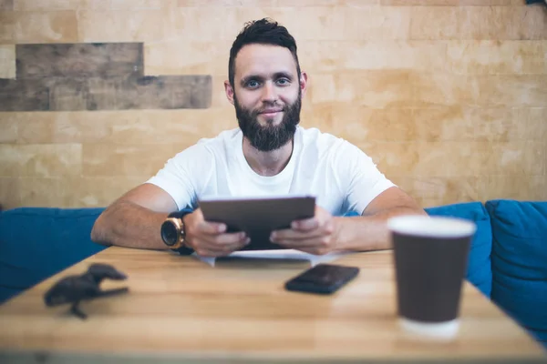 Hombre usando una tableta PC en un restaurante o cafetería. Lleva una camiseta blanca y está sentado junto a una mesa. —  Fotos de Stock