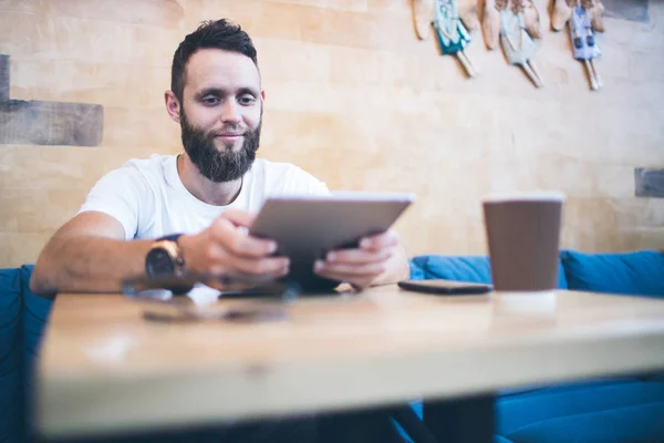 Hombre usando una tableta PC en un restaurante o cafetería. Lleva una camiseta blanca y está sentado junto a una mesa. —  Fotos de Stock