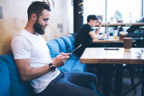 Homme utilisant une tablette PC dans un restaurant ou un café. Il porte un t-shirt blanc et est assis près d'une table. — Photo
