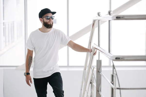 Hipster beau modèle masculin avec barbe portant un t-shirt blanc et une casquette de baseball avec de l'espace pour votre logo ou design dans un style urbain décontracté — Photo