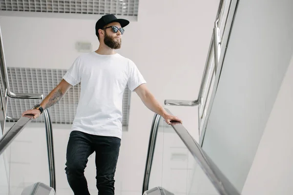 Hipster όμορφο αρσενικό μοντέλο με γενειάδα φορώντας λευκό κενό t-shirt και ένα καπέλο του μπέιζμπολ με χώρο για το λογότυπο ή το σχέδιό σας σε casual αστικό στυλ — Φωτογραφία Αρχείου