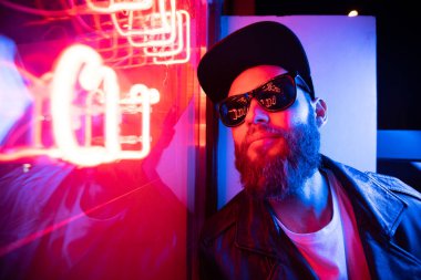 Şehir sokaklarında hipster yakışıklı adam Neon işaretler tarafından aydınlatılmış olmak. O siyah kap, kot pantolon ve güneş gözlüğü ile deri motosikletçi ceket veya asimetrik fermuar ceket giyiyor.