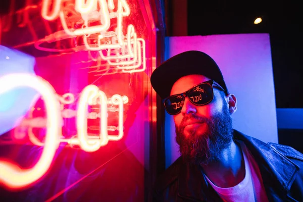 Hipster hombre guapo en las calles de la ciudad siendo iluminado por carteles de neón. Lleva chaqueta motorista de cuero o chaqueta con cremallera asimétrica con gorra negra, jeans y gafas de sol. . — Foto de Stock