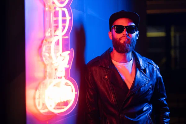 Hipster gutaussehender Mann auf den Straßen der Stadt, beleuchtet von Leuchtreklamen. er trägt Biker-Lederjacke oder asymmetrische Reißverschlussjacke mit schwarzer Mütze, Jeans und Sonnenbrille. — Stockfoto