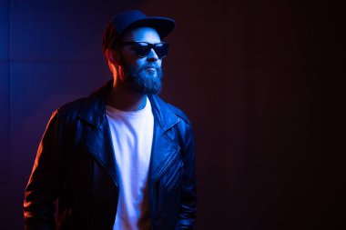 Şehir sokaklarında hipster yakışıklı adam Neon işaretler tarafından aydınlatılmış olmak. O siyah kap, kot pantolon ve güneş gözlüğü ile deri motosikletçi ceket veya asimetrik fermuar ceket giyiyor.