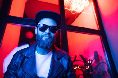 Şehir sokaklarında hipster yakışıklı adam Neon işaretler tarafından aydınlatılmış olmak. O siyah kap, kot pantolon ve güneş gözlüğü ile deri motosikletçi ceket veya asimetrik fermuar ceket giyiyor.
