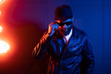 Şehir sokaklarında hipster yakışıklı adam Neon işaretler tarafından aydınlatılmış olmak. O siyah kap, kot pantolon ve güneş gözlüğü ile deri motosikletçi ceket veya asimetrik fermuar ceket giyiyor.
