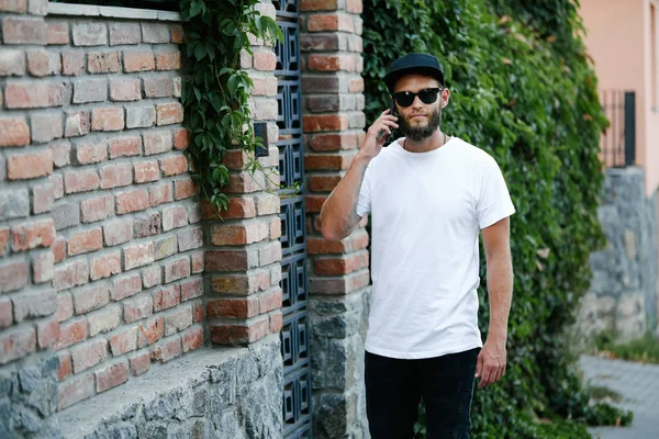 Modelo masculino bonito Hipster com barba vestindo t-shirt branca em branco e um boné de beisebol com espaço para o seu logotipo ou design em estilo urbano casual — Fotografia de Stock