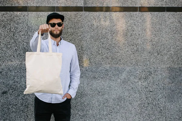 Jovem segurando saco eco têxtil branco contra fundo urbano da cidade. .. Conceito de ecologia ou de protecção do ambiente. Saco eco branco para simular . — Fotografia de Stock