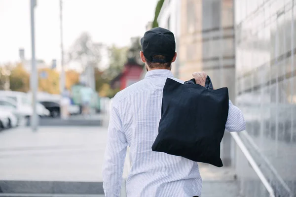 Jonge man Holding zwarte textiel Eco tas tegen stedelijke stad achtergrond. . Ecologie of milieubescherming concept. Zwarte Eco-tas voor uw ontwerp of logo mock up — Stockfoto