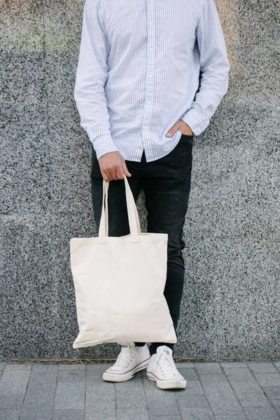 Junger Mann mit weißer Textil-Öko-Tasche vor städtischem Hintergrund. . Ökologie oder Umweltschutzkonzept. weiße Öko-Tasche für Attrappen. — Stockfoto