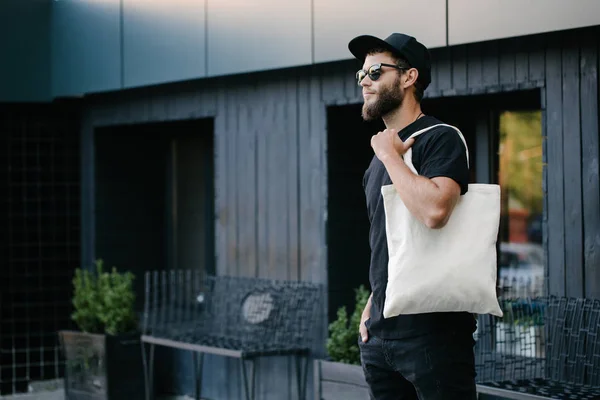 Ung man innehar vit textil Eco Bag mot Urban City bakgrund. . Ekologin eller miljöskydds konceptet. Vit Eco Bag för mock-up. — Stockfoto