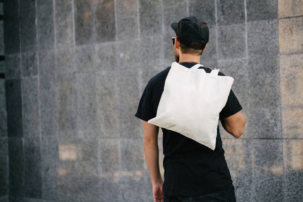 Молодой человек держит белую текстильную эко-сумку на фоне города. - Да. Концепция экологии или охраны окружающей среды. Белая экосумка для насмешек
.