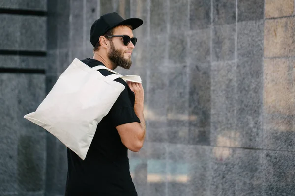 Νέος άνθρωπος που κατέχει λευκό υφαντουργική τσάντα Eco κατά του αστικού ιστορικού της πόλης. . Οικολογία ή έννοια προστασίας του περιβάλλοντος. Λευκή τσάντα Eco για την αναπαράσταση. — Φωτογραφία Αρχείου