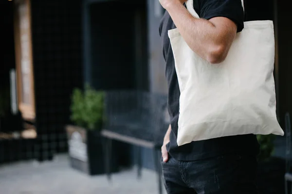 Ung man innehar vit textil Eco Bag mot Urban City bakgrund. . Ekologin eller miljöskydds konceptet. Vit Eco Bag för mock-up. — Stockfoto
