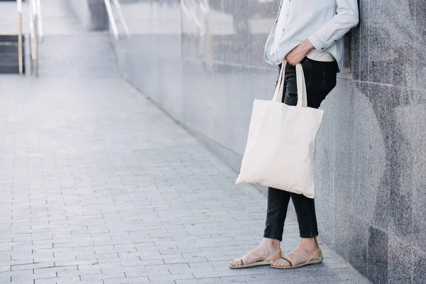 Mulher segurando saco eco têxtil branco contra fundo urbano da cidade. .. Conceito de ecologia ou de protecção do ambiente. Saco eco branco para simular . — Fotografia de Stock