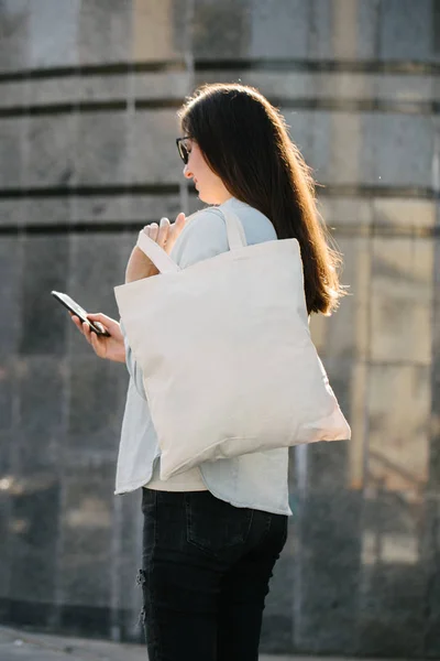 Frau mit weißer Textil-Öko-Tasche vor städtischem Hintergrund. . Ökologie oder Umweltschutzkonzept. weiße Öko-Tasche für Attrappen. — Stockfoto