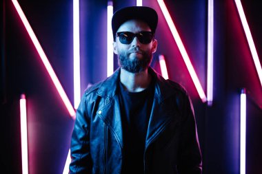 Şehir sokaklarında hipster yakışıklı adam Neon işaretler tarafından aydınlatılmış olmak. O siyah kap, kot pantolon ve güneş gözlüğü ile deri motosikletçi ceket veya asimetrik fermuar ceket giyiyor.
