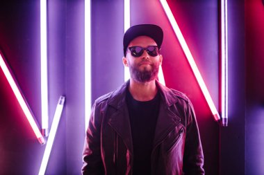 Şehir sokaklarında hipster yakışıklı adam Neon işaretler tarafından aydınlatılmış olmak. O siyah kap, kot pantolon ve güneş gözlüğü ile deri motosikletçi ceket veya asimetrik fermuar ceket giyiyor.