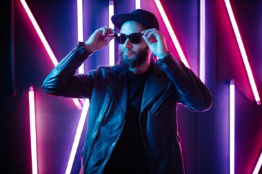 Şehir sokaklarında hipster yakışıklı adam Neon işaretler tarafından aydınlatılmış olmak. O siyah kap, kot pantolon ve güneş gözlüğü ile deri motosikletçi ceket veya asimetrik fermuar ceket giyiyor.