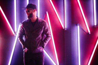 Şehir sokaklarında hipster yakışıklı adam Neon işaretler tarafından aydınlatılmış olmak. O siyah kap, kot pantolon ve güneş gözlüğü ile deri motosikletçi ceket veya asimetrik fermuar ceket giyiyor.
