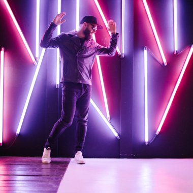 Şehir sokaklarında hipster yakışıklı adam Neon işaretler tarafından aydınlatılmış olmak. O siyah kap, kot pantolon ve güneş gözlüğü ile deri motosikletçi ceket veya asimetrik fermuar ceket giyiyor.