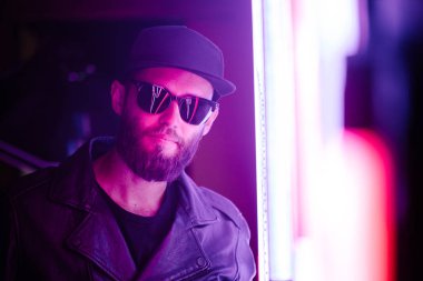 Şehir sokaklarında hipster yakışıklı adam Neon işaretler tarafından aydınlatılmış olmak. O siyah kap, kot pantolon ve güneş gözlüğü ile deri motosikletçi ceket veya asimetrik fermuar ceket giyiyor.