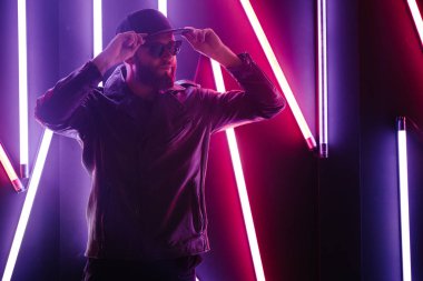 Şehir sokaklarında hipster yakışıklı adam Neon işaretler tarafından aydınlatılmış olmak. O siyah kap, kot pantolon ve güneş gözlüğü ile deri motosikletçi ceket veya asimetrik fermuar ceket giyiyor.